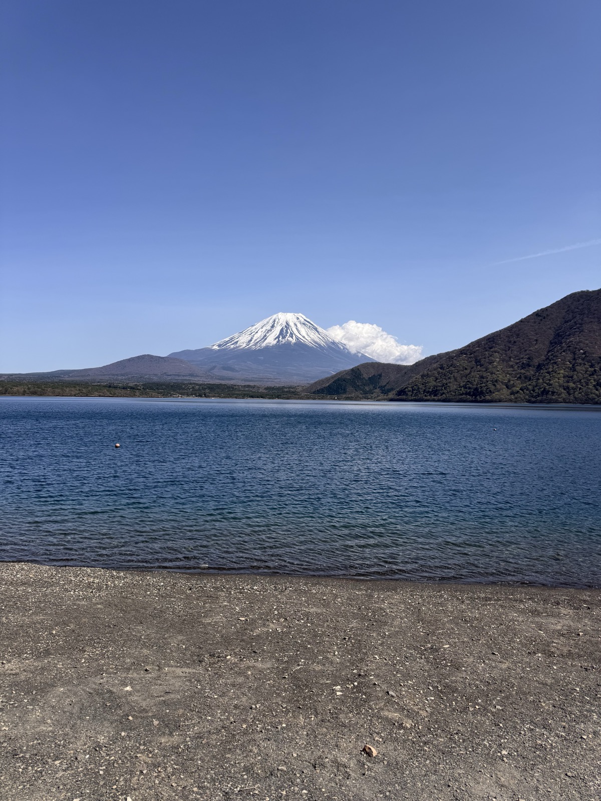 Snow-capped Mount Fuji rising beyond Lake Motosu, seen from the pebble shore at a lakeside campsite